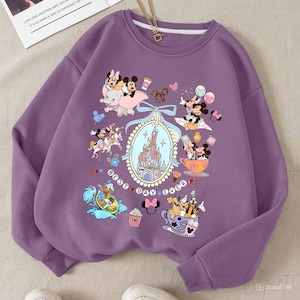 May include: A purple sweatshirt featuring a colorful Disney-themed graphic. The design includes Mickey and Minnie Mouse, a castle, and the text "Best Day Ever." The sweatshirt has a crew neck and long sleeves.