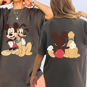May include: Dark gray t-shirt featuring a cartoon illustration of Mickey Mouse and Pluto. Mickey is wearing red shorts and white gloves, Pluto has a green collar. The back of the shirt shows the same characters.