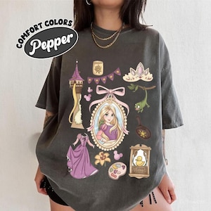 May include: A dark gray t-shirt with a graphic design featuring a princess, a tower, and other related elements. The shirt has the words "Comfort Colors" and "Pepper" printed on it.