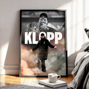 Könnte beinhalten: Schwarz gerahmtes Poster mit einem schwarz-weiß Bild eines Mannes, der eine Trophäe hält, mit dem Wort "KLOPP" in Weiß. Ein zweites Bild des Mannes ist darüber gelegt. Auf dem Boden liegt ein Buch und eine Tasse Kaffee.