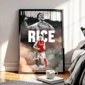 May include: A framed poster featuring a soccer player in a red and white jersey, with the name "RICE" in large white letters. The image has a smoky background and a black frame. A coffee cup sits on a book in the foreground.