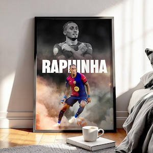 Puede incluir: Un póster enmarcado con un futbolista con uniforme rojo, azul y amarillo, con el nombre "RAPHINHA" en letras blancas grandes. El fondo tiene un efecto ahumado, y una taza de café está sobre un libro en primer plano.