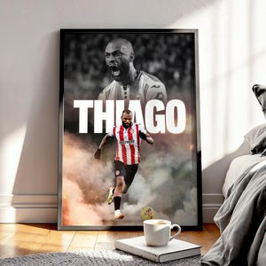 May include: A framed poster with a football player in a red and white striped jersey, with the name "THIAGO" in white. The background is black and white. A football is near the player's feet. The poster is displayed on a wooden floor.