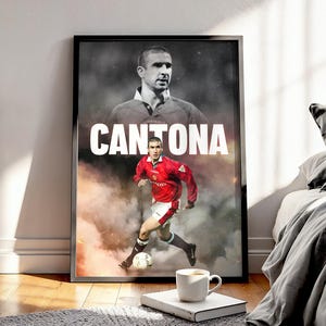Op de afbeelding: Een ingelijste poster met een voetballer in een rood shirt en witte shorts, die tegen een voetbal trapt. De poster heeft de naam "CANTONA" in grote witte letters. Een tweede afbeelding van de speler staat op de achtergrond.