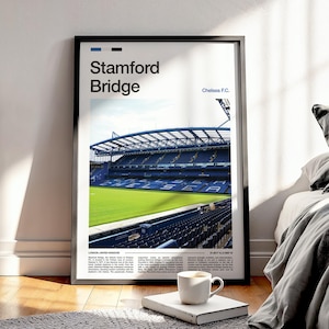 Puede incluir: Un póster enmarcado con el Stamford Bridge, el estadio del Chelsea F.C. La imagen muestra los asientos azules del estadio, el campo verde y el texto "Stamford Bridge" y "Chelsea F.C." en blanco. Una taza de café descansa sobre un libro frente al póster.