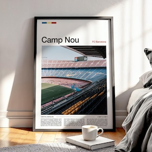Op de afbeelding: Een ingelijste poster met het Camp Nou-stadion, de thuisbasis van FC Barcelona, met de tekst "Camp Nou" en "FC Barcelona". De afbeelding toont de binnenkant van het stadion met blauwe, rode en gele stoelen en een groen veld. Een koffiekopje staat op een boek voor de poster.