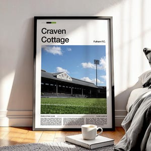 May include: Framed poster featuring Craven Cottage stadium with the text "Craven Cottage" and "Fulham F.C." against a blue sky. The image shows the stadium's exterior, green field, and a floodlight. A white mug sits on a book in front of the poster.