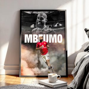Könnte beinhalten: Gerahmtes Poster mit einem Fußballspieler in einem roten Trikot und dem Namen "MBEUMO" in Weiß. Das Bild enthält ein Schwarz-Weiß-Foto des Spielers über dem Farbfoto, mit einer Kaffeetasse und einem Buch im Vordergrund.