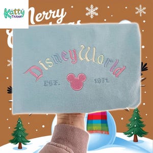 A hand holding a light blue item with the text "DisneyWorld" and "Est. 1971" embroidered on it, against a background of snowflakes and Christmas trees.