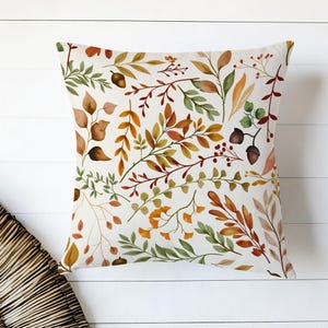 May include: Square throw pillow with a white background and a watercolor pattern of autumn leaves and acorns in shades of green, orange, and brown. The pillow is likely made of a soft fabric and is designed for decorative use.