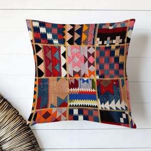 Puede incluir: Cojín cuadrado con diseño de patchwork. El cojín presenta una variedad de patrones geométricos en tonos de rojo, azul, naranja, rosa y beige. El cojín está colocado sobre un fondo de madera blanca.
