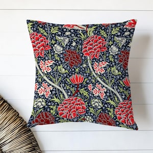 May include: Square throw pillow with a floral pattern. The pillow features a dark blue background with a repeating design of red and white flowers and green leaves. The pillow is placed on a white wooden surface.