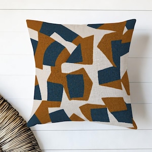 May include: Square throw pillow with a geometric pattern in navy blue and rust-colored shapes on a cream background. The pillow has a textured appearance and is likely made of fabric.