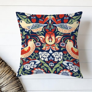 Op de afbeelding: Vierkant decoratief kussen met een marineblauwe achtergrond en een herhalend patroon van vogels, bloemen en bladeren in rood, crème en groen. De vogels hebben rode en oranje markeringen. Het kussen ligt op een wit houten oppervlak.