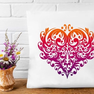 Peut inclure: Coussin carré blanc avec un grand motif de cœur orné. Le cœur présente un dégradé de couleurs, passant de l'orange en haut au violet foncé en bas. Un vase de fleurs est placé à gauche du coussin.