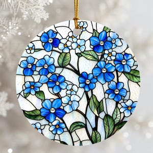 May include: A round, ceramic ornament featuring a stained-glass design of blue flowers with green leaves. The flowers have jewel-like centers. The ornament is suspended by a gold cord, perfect for holiday decor.