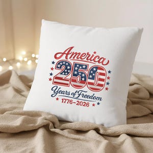 May include: A white square cushion featuring the embroidered text "America 250 Years of Freedom 1776-2026". The design uses red, white, and blue, with stars and stripes, commemorating the anniversary.