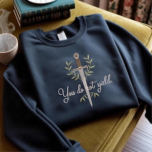 Op de afbeelding: Marineblauw sweatshirt met een geborduurd zwaardontwerp, omringd door olijftakken, en de tekst "You do not yield". Het sweatshirt wordt getoond op een geelgroen kussen, met boeken en een mok op de achtergrond.