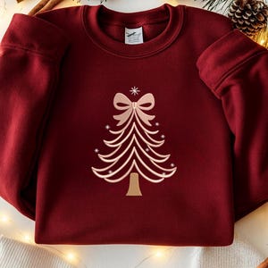 May include: A burgundy sweatshirt featuring a light pink embroidered Christmas tree design. The tree has a bow at the top and small star accents. The sweatshirt has a crew neck and long sleeves.