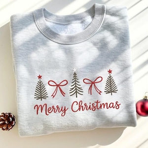 May include: A light gray sweatshirt with embroidered Christmas trees, red bows, and the words "Merry Christmas" in red script. The design includes a star on top of each tree. A pinecone and a red ornament are in the background.