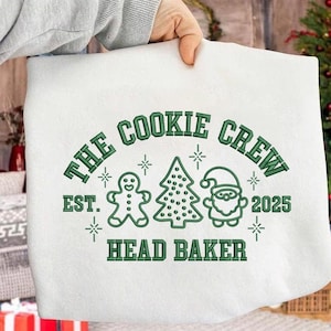 Peut inclure: Sweat-shirt blanc avec un motif vert. Le motif comprend le texte "THE COOKIE CREW", un bonhomme en pain d'épices, un sapin de Noël et le Père Noël. Le texte "EST. 2025 HEAD BAKER" est également inclus.