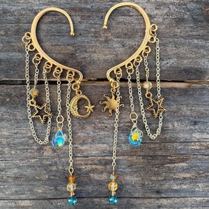 May include: A pair of gold-toned ear cuffs featuring dangling chains, star charms, a crescent moon charm, and a sun charm. The jewelry includes small faceted beads in blue, yellow, and clear tones attached to the ends of the delicate metal chains.