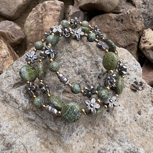 May include: A multi-strand wrap bracelet featuring round, textured green stone beads interspersed with small, metallic gold-toned spacer beads and silver-toned flower-shaped charms. The bracelet is constructed with a flexible wire design.