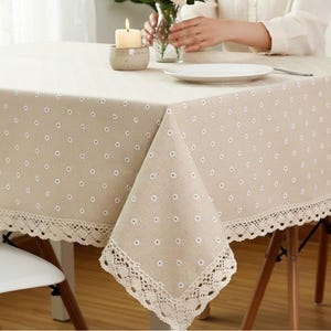 Peut inclure: Une nappe beige ornée d'un motif répétitif de petites fleurs de marguerite blanches aux cœurs jaunes. Les bords du tissu sont finis avec une large bordure en dentelle au crochet de couleur crème qui pend le long de la surface de la table.