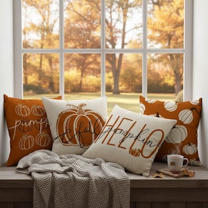 May include: Three square scatter cushions featuring autumn themes. One is burnt orange with white line-art pumpkins, one is cream with a large orange pumpkin and the text "fall", and one is cream with the text "HELLO pumpkin" in orange script.