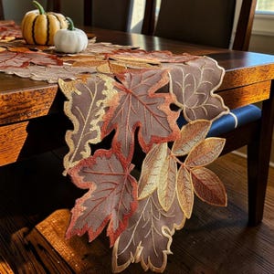 May include: A table runner featuring an intricate die-cut design of autumn leaves in shades of brown, orange, and gold. The fabric leaves are embroidered with detailed veins and scalloped edges, creating a layered, textured appearance on the table.