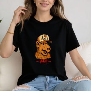 May include: Black t-shirt featuring a cartoon character wearing a hat that says "I ❤️ CATS." The character has orange fur and a large nose. The text "EST. ALF 1986" is printed in red below the character.