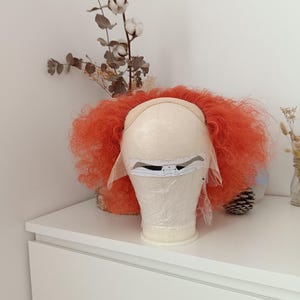 May include: A bright orange curly wig on a mannequin head. The wig features a lace front cap with a white paper measuring guide attached across the forehead area. The hair is voluminous with tight curls, extending outward from the center.