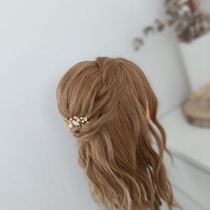 Peut inclure: Une tête de mannequin avec des cheveux ondulés brun clair coiffés avec une barrette dorée et perles. La barrette est positionnée sur le côté de la tête, ajoutant une touche d'élégance. Le fond est un mur blanc uni.