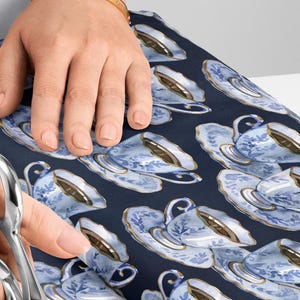 Pode incluir: Uma pessoa corta tecido com uma tesoura. O tecido tem um padrão repetido de xícaras de chá azuis e brancas sobre um fundo azul escuro. Uma régua de madeira é visível. A pessoa usa um top branco e uma pulseira dourada.