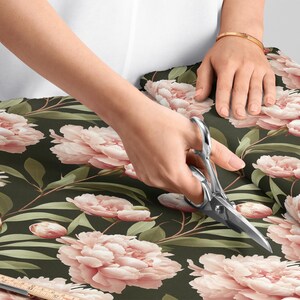 Puede incluir: Una persona usa tijeras para cortar tela con un estampado floral. La tela presenta grandes flores de peonías rosas y hojas verdes sobre un fondo verde oscuro. Se ve una regla de madera.