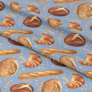 Pode incluir: Tecido azul claro com um padrão repetido de vários produtos de panificação, incluindo croissants, baguetes e pães redondos. As ilustrações em estilo aquarela apresentam tons de castanho e bege, com espigas de trigo.