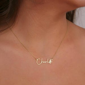 Könnte beinhalten: Eine zarte, goldfarbene Halskette mit dem Namen "Charlotte" in Schreibschrift. Der Anhänger hängt an einer feinen Kette und erzeugt ein minimalistisches und elegantes Design. Die Halskette wird vor einem hautfarbenen Hintergrund gezeigt.