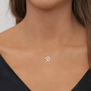 May include: A gold necklace featuring a fine chain and a cursive letter "D" pendant. The pendant hangs centrally, offering a simple, elegant aesthetic. The necklace is displayed against a dark backdrop.
