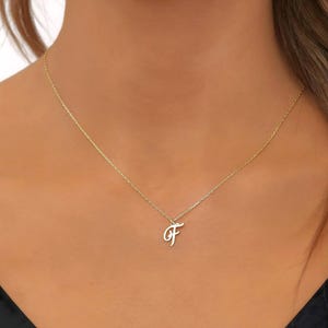 Könnte beinhalten: Eine zarte, goldfarbene Halskette mit einem Anhänger in Form eines kursiven Buchstaben "F". Der Anhänger hängt an einer feinen Kette. Ein schlichtes, elegantes Schmuckstück.