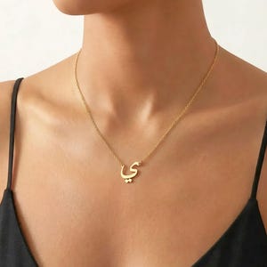 Peut inclure: Un collier pendentif de couleur or avec une lettre arabe. La chaîne délicate suspend la lettre stylisée, qui est le point central du collier. Le collier est porté sur un fond sombre.