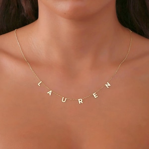 Peut inclure: Un délicat collier en or épelant le nom "LAUREN" en lettres majuscules individuelles. Les lettres sont suspendues à une fine chaîne, créant un accessoire personnalisé et élégant. Le collier est présenté sur un fond de teint.