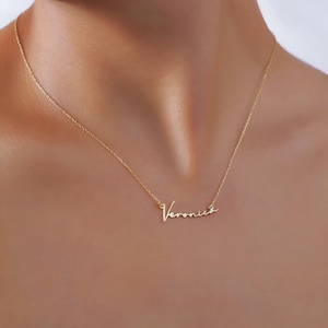 May include: A delicate gold-colored necklace with the name "Veronica" in a cursive script. The necklace is worn around the neck, with the chain and pendant visible against the skin.