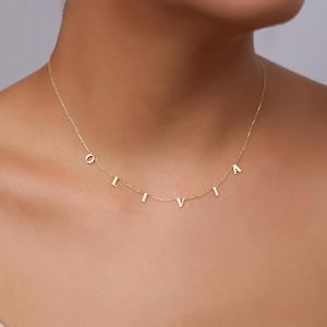 Op de afbeelding: Een delicate gouden ketting met de naam "OLIVIA" in afzonderlijke hoofdletters. De letters zijn aan een fijne ketting bevestigd, waardoor een gepersonaliseerd en elegant accessoire ontstaat. De ketting wordt om de nek van een persoon getoond.