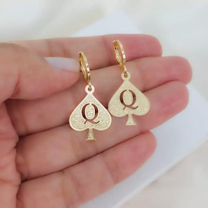 May include: A pair of gold-toned hoop earrings featuring a dangling spade-shaped charm. Each charm has a textured surface and a cutout letter Q in the centre. The earrings are designed with a hinged closure mechanism for secure attachment to the ear.