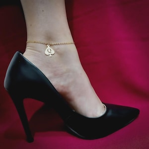 May include: Gold-colored anklet with a spade charm and the letter "Q". The anklet is worn with a black high-heeled shoe. The background is a solid, deep red color.