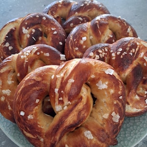 May include: A selection of freshly baked pretzels, presented on a light green plate. The pretzels are a rich golden brown with darker, caramelised patches and sprinkled with coarse salt crystals. A classic baked treat.