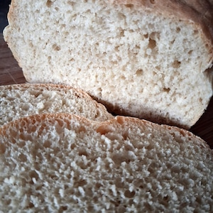 May include: A loaf of freshly baked bread, partially sliced, with a light brown crust and a porous interior. The bread has a rustic appearance, with visible air pockets and a soft texture. The slices are cut to a medium thickness, ready for serving.