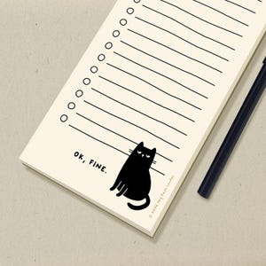 May include: A cream-coloured notepad featuring a black cat illustration and the words "OK, FINE." The notepad has lines and circles for checklists. A black pen lies beside it. The notepad is from Hey Rose London.
