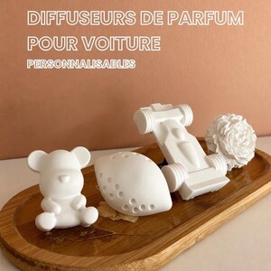 Peut inclure: Quatre diffuseurs de parfum pour voiture blancs mats en forme d'ours en peluche, de citron, de voiture de course et de fleur, disposés sur un plateau ovale en bois. Le texte DIFFUSEURS DE PARFUM POUR VOITURE PERSONNALISABLES est visible.