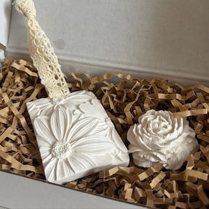 Peut inclure: Un porte-savon rectangulaire blanc avec un motif de marguerites et une poignée en dentelle crème. Un savon blanc en forme de rose est posé à côté, niché dans du rembourrage en papier brun à l'intérieur d'une boîte. Articles de décoration.